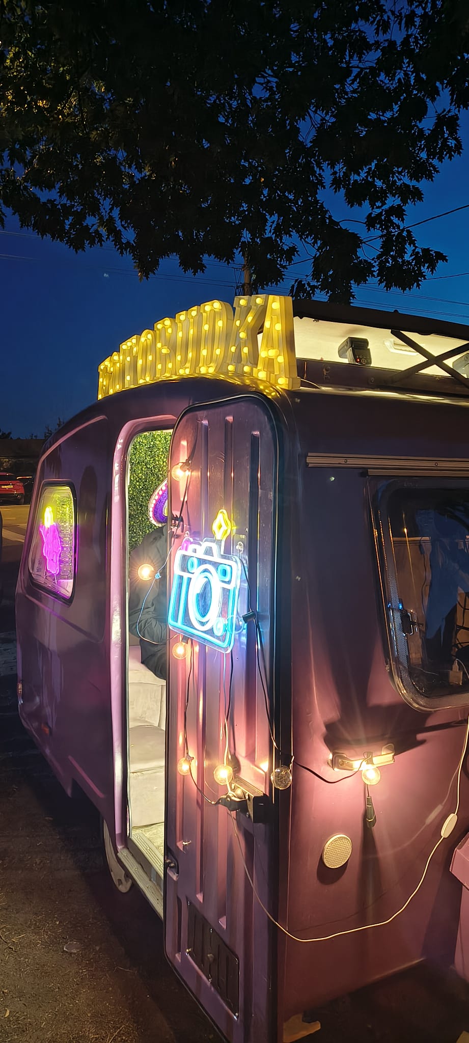 Fioletowa fotoprzyczepa Fotofurka wieczorem, podświetlony neon aparatu i szyld z napisem